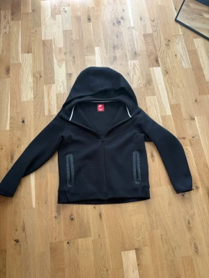 Nike tech  - Säljer en svart hoodie från Jd med hel dragkedja framtill och två fickor med dragkedja. Hoodien har en stor huva och diskret Nike-logga på bröstet. 