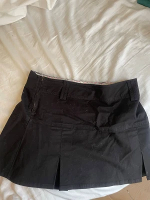 Svart kortkjol med blommigt foder - Säljer en svart kortkjol med fina veck och bälteshällor. Insidan har ett gulligt blommigt foder i rosa och grönt. 