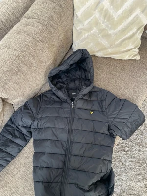 Svart dunjacka från Lyle & Scott - Snygg svart dunjacka från Lyle & Scott Junior med huva och dragkedja framtill. Jackan har ett quiltat mönster och den klassiska Lyle & Scott-loggan i gult på bröstet. Perfekt för kalla vinterdagar och riktigt stilren.