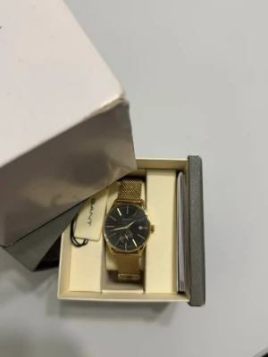 Guld klocka med svart urtavla från Gant - Stilren klocka från Gant med guldfärgat mesh-armband och svart urtavla. Klockan har minimalistiska guldfärgade markeringar och visare. Levereras i originalask med tillhörande manual. Perfekt accessoar för dig som gillar klassisk och elegant stil.