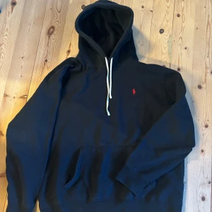 Svart hoodie från Polo Ralph Lauren - Svart hoodie från Polo Ralph Lauren med klassisk röd logga broderad på bröstet. Hoodien har vit dragsko i huvan och en stor magficka framtill. Tillverkad i mjukt bomullsmaterial och har en avslappnad passform.