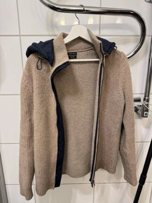 Beige stickad jacka Massimo Dutti - Stickad beige jacka från Massimo Dutti i extrafin ull med mörkblå quiltad huva. Jackan har dragkedja framtill, ribbade muddar och långärmad design. Perfekt för dig som gillar chill och stilren look med en twist av sportig känsla. Storleken är Mellan S och M så den kommer passa dig utmärkt om du har en av dessa storlekarna