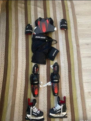 Svart bauer hockey kläder som är hel - Säljer en komplett hockeyutrustning från Bauer med svarta och röda skydd, axelskydd, armbågsskydd, benskydd, hockeybyxor och handskar. Allt är designat för att ge maximalt skydd och komfort på isen. Materialet är slitstarkt och anpassat för hockey.