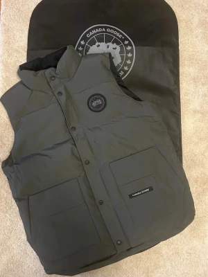 Grå dunväst från Canada Goose - Snygg grå dunväst från Canada Goose med hög krage, två stora framfickor och svart logomärke på bröstet. Västen har tryckknappar framtill och är tillverkad i slitstarkt material med dunfyllning för extra värme.