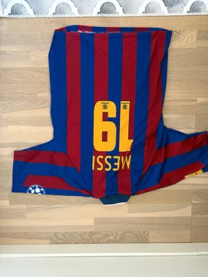 Barcelona Messi matchtröja röd/blå - Säljer en klassisk Barcelona matchtröja med Messi och nummer 19 på ryggen. Tröjan har vertikala röda och blå ränder, klubbmärke på bröstet och Champions League-märke på ärmen. Tillverkad i lätt och ventilerande material, perfekt för fotboll eller samling.