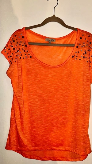Färgstark orange topp med dekorativa stenar – Sweet Miss  - Säljer en jättefin topp från märket Sweet Miss i en härlig, klarorange nyans. Toppen har en smickrande rund halsringning som är vackert dekorerad med svarta stenar och paljetter, vilket ger en snygg kontrast och en festlig känsla. Modellen är rymlig och har en skön, något "oversized" passform. Materialet är mjukt och faller fint. Mått: Bredd ca 57 cm Längd ca 70 cm