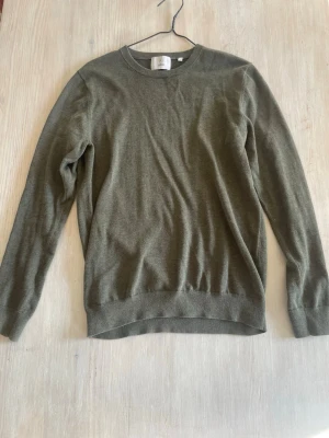 Olivgrön stickad tröja från Bläck - Olivgrön stickad tröja från Bläck med rund halsringning och ribbade muddar vid ärmslut och nederkant. Enkel och stilren design som passar perfekt till lager-på-lager. Tillverkad i mjukt material för skön känsla.