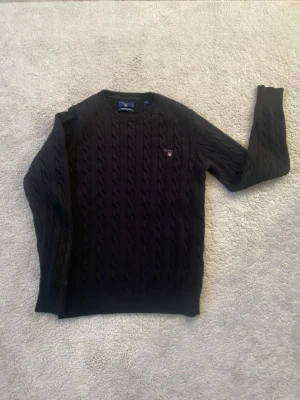 Svart kabelstickad tröja från Gant - Säljer en svart kabelstickad tröja från Gant med rund halsringning och diskret logga på bröstet. Tröjan har långa ärmar och ribbade muddar vid ärmslut och nederkant. Perfekt för dig som gillar klassisk och stilren design.