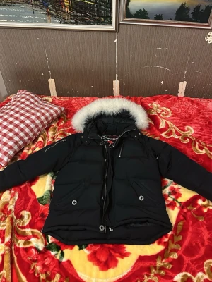 Svart dunjacka från moose knuckles  - Snygg svart dunjacka med stor huva som har en fluffig, vit pälskant. Jackan har dragkedja och knappar framtill, två framfickor med knapp och quiltad design. Perfekt för kalla vinterdagar och riktigt varm. Passar dig som vill ha både stil och funktion.