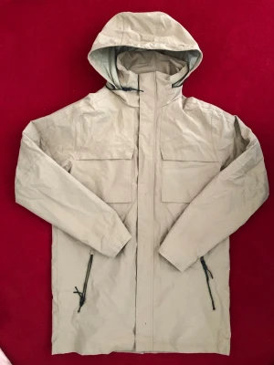 Mckinley Beige regnjacka med huva - Stilren beige regnjacka med huva och dragkedja framtill. Jackan har två stora bröstfickor med lock och två snedställda fickor med dragkedja. Tillverkad i vattenavvisande material och har justerbar huva för extra skydd mot regn.