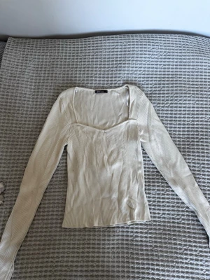 Ribbad beige topp från Gina Tricot - Trendig ribbad topp från Gina Tricot i beige med lång ärm och snygg fyrkantig urringning. Toppen har en figurnära passform och är tillverkad i mjuk bomullsmix som känns skön mot huden. Perfekt att styla med jeans eller kjol för en clean look.