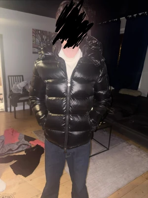 Svart glansig pufferjacka med huva - Säljer en svart, glansig pufferjacka med stor huva och dragkedja framtill. Jackan har breda kanaler och två sidofickor. Perfekt för kalla vinterdagar och ger en riktigt modern look. 