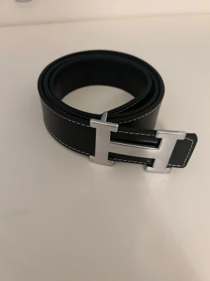 Svart  läderbälte med H-spänne - Snyggt svart bälte från i äkta läder med kontrasterande vita sömmar. Bältet har ett ikoniskt H-format metallspänne i silverfärg som ger en exklusiv känsla. Perfekt accessoar för att lyfta vilken outfit som helst.