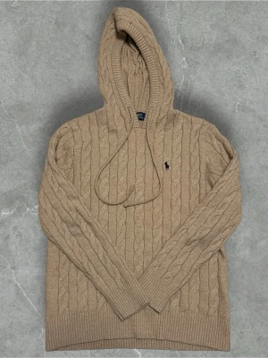 Beige kabelstickad hoodie Polo Ralph Lauren i 100% kashmir - Säljer en beige kabelstickad hoodie från Polo Ralph Lauren med klassisk broderad logga på bröstet. Tröjan har huva med dragsko, långa ärmar och ribbade muddar. Perfekt för en avslappnad och stilren look. Passar S och m