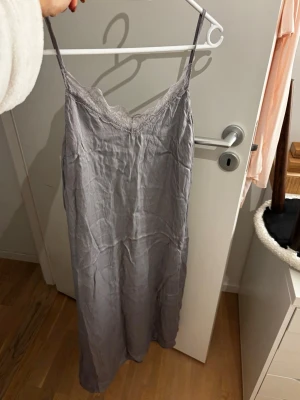 Ljusgrå nattlinne med spetsdetaljer - Säljer ett ljusgråa nattlinne med tunna axelband och vacker spets längs urringningen. Nattlinnet har en rak och avslappnad passform, perfekt för mysiga kvällar hemma. Materialet känns mjukt mot huden och ger en lyxig känsla. Den är aldrig använd men ser skrynklig ut pga att jag tvättat den. Storlek M men passar en S. 