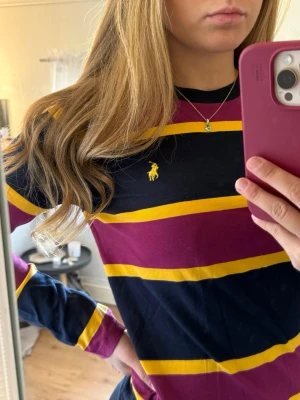 Randig långärmad topp från Ralph Lauren - Säljer en långärmad topp från Ralph Lauren med breda marinblå, vinröda och gula ränder. Klassisk gul logga broderad på bröstet. Skön bomullskvalitet och normal passform. Perfekt för dig som gillar sportig och stilren look.