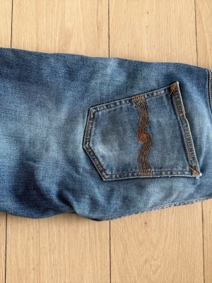 Blå slitna jeans från Nudie Jeans - Ett par blå jeans från Nudie Jeans med slitningar och hål på benen för en riktigt avslappnad look. Klassisk femficksmodell med Nudie Jeans signaturbrodyr på bakfickan. Tillverkade i denim med snygg tvätt och raka ben.