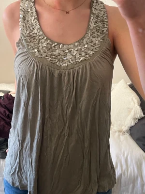 Grönt linne med paljetter - Snyggt grönt linne med rundad hals och stora silverfärgade paljetter längs urringningen. Luftigt material och avslappnad passform.