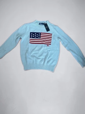 Ljusblå stickad tröja Polo Ralph Lauren - Ljusblå stickad tröja från Polo Ralph Lauren med amerikansk flagga och RL-broderi på bröstet. Tröjan har rund halsringning och ribbstickade muddar vid ärmslut och nederkant. Perfekt för dig som gillar klassisk och preppy stil.