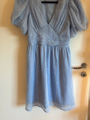 Ljusblå kortklänning med puffärm - Supersöt ljusblå kortklänning med puffiga ärmar och v-ringning. Klänningen har ett diskret vitt mönster, markerad midja och lätt rynkade detaljer framtill. Perfekt för sommarens alla tillfällen!