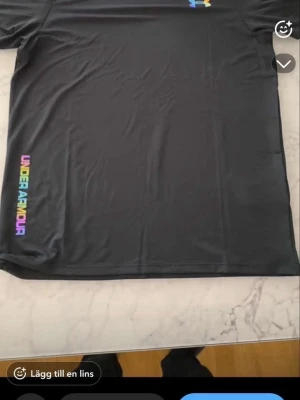 Svart träningsset från Under Armour - Snyggt svart träningsset från Under Armour med regnbågsfärgade logotyper. Setet består av en t-shirt och shorts i lätt och snabbtorkande material, perfekt för träning eller sport. Både tröjan och shortsen har Under Armour-tryck i färg på sidan.