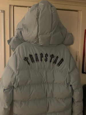Trapstar Ljusblå Puffer Jacket - Snygg ljusblå pufferjacka från Trapstar med huva och dragkedja framtill. Jackan har ett runt emblem på bröstet och är quiltad för extra värme. Perfekt för kalla dagar och riktigt trendig look.