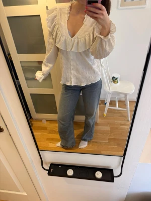 Vit volangblus från Monki - Supersöt vit blus från Monki med stora volanger längs ringningen och v-ringad hals. Blusen har långärmade puffärmar med resår vid ärmslut och är gjord i ett strukturerat, lätt tyg. Perfekt för en trendig look. Väl använd, men inga deffekter! 