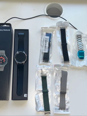 Samsung Galaxy Watch5 med extra armband - Samsung Galaxy Watch5 smartklocka i svart med rund urtavla och metallram. Medföljer flera utbytbara armband i olika färger och material: svart mesh, grönt tyg, grått silikon, blått silikon och ett silverfärgat metallarmband. Perfekt för dig som vill variera stilen.