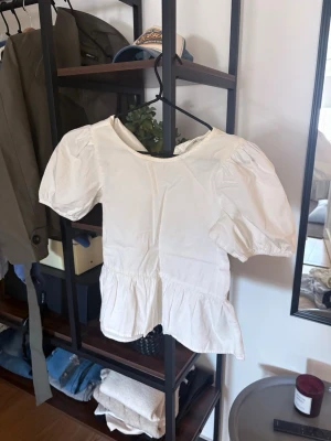 Vit puffärm blus med volang - Söt vit blus med puffiga korta ärmar och volang nedtill. Blusen har rund halsringning och är tillverkad i ett lätt tyg som ger en luftig känsla. Perfekt för en trendig och avslappnad stil.