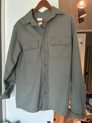 Mörkgrön overshirt från H&M - Stilren mörkgrön overshirt från H&M med raka linjer och två stora bröstfickor med lock och knapp. Skjortan har klassisk krage, knäppning framtill och är tillverkad i ett slitstarkt bomullstyg. Perfekt lager-på-lager-plagg för en avslappnad look.