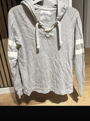 Grå hoodie från  - Snygg grå hoodie från Iconic Sport med vita snören och cool snörning vid halsen. Tröjan har vita ränder på ärmarna och en klassisk huva. Tillverkad i mjukt bomullsmaterial som känns skönt mot huden. Perfekt för en avslappnad och sportig stil.