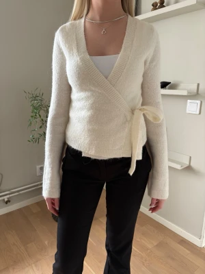 Vit omlottstickad tröja med knyt - Mysig vit stickad omlottopp med knyt i sidan och v-ringad hals. Tröjan har lång ärm och ribbade muddar. Perfekt att styla över ett linne eller topp för en enkel men snygg look.