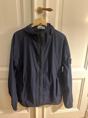 Stone Island vindjacka - Snygg blå vindjacka från Stone Island med huva och dragkedja framtill. Jackan har två sidofickor med dragkedja och klassisk logotyp på vänster ärm. Tillverkad i lätt och slitstarkt material som passar perfekt för blåsiga dagar