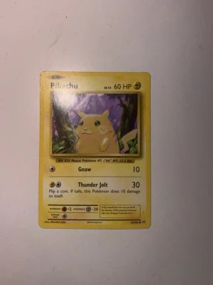 Pikachu Pokémonkort Base Set - Säljer ett klassiskt Pikachu-kort från Pokémon Base Set. Kortet är guldfärgat med motiv av Pikachu i en skogsmiljö, har 60 HP och attacker som Gnaw och Thunder Jolt. Kortet är tryckt på tjockt papper och har detaljerad text och symboler. Buda på