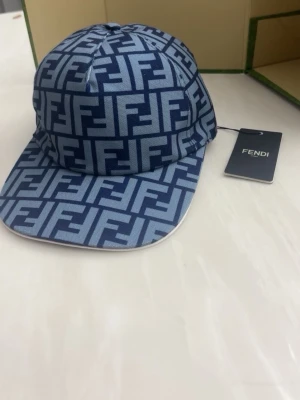 Marin blå FF keps - Säljer en blå keps från FF med ikoniskt FF-mönster i två nyanser av blått. Kepsen har böjd skärm och justerbar baksida. Tillverkad i ett slitstarkt tyg som ger en exklusiv känsla. Perfekt för dig som vill sticka ut med en lyxig accessoar.