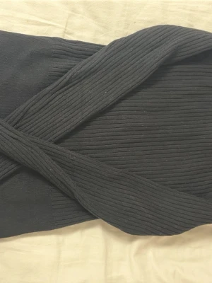 Svart ribbad omlottopp - Snygg svart omlottopp med ribbad struktur och breda axelband som korsas framtill. Perfekt för dig som gillar en stilren och trendig look. Toppen är ärmlös och har en tight passform som framhäver figuren.