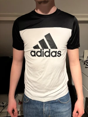 Svartvit Adidas t-shirt med logga - Snygg svartvit t-shirt från Adidas med stor logga framtill. T-shirten har korta ärmar, rund halsringning och är tillverkad i ett lätt och funktionellt material som andas. Perfekt för dig som gillar sportig stil och vill ha något bekvämt.