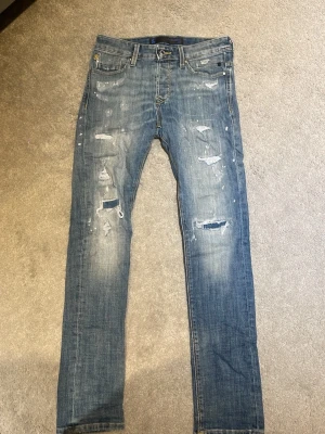 Blå slitna jeans från jacknjones  - Säljer ett par blå jeans från jacknjones med skinny fit och slitningar framtill. Jeansen har distressed detaljer, hål och färgstänk för en cool, trendig look. Klassisk femficksmodell med knappgylf och patch bak i midjan. Perfekta för dig som gillar streetstyle.