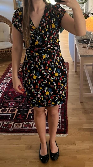 Moschino klänning - Kort Moschino klänning med färgglatt blommönster i gult, blått, rött och vitt på svart botten. Klänningen har v-ringning, knyt i midjan och korta ärmar. Perfekt för dig som gillar färg och mönster!