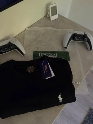 Svart t-shirt från Polo Ralph Lauren - Klassisk svart t-shirt från Polo Ralph Lauren med rund halsringning och ikoniska vita logobroderiet på bröstet. Tillverkad i mjuk bomull för en skön känsla och perfekt passform. En stilren basic som funkar till allt.