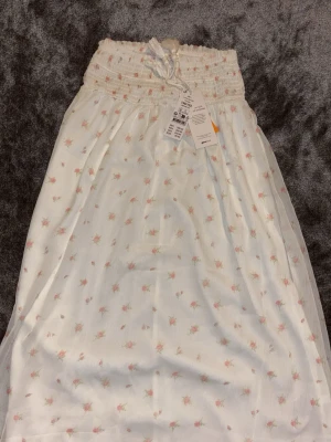 Två långa kjolar med mönster - Säljer två långa kjolar med resår och knytband i midjan. Ena kjolen är vit med små blommor i rosa och grönt, den andra är ljusblå med diskret paisleymönster. Båda har en luftig och flowy känsla, perfekta för varma dagar.