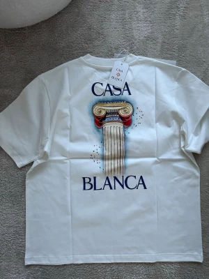 Vit Casablanca t-shirt med pelartryck - Vit t-shirt från Casablanca med stort tryck av en klassisk pelare och texten 'CASA BLANCA' på ryggen samt mindre tryck på bröstet. T-shirten har rund hals och korta ärmar. Tillverkad i mjuk bomull för en clean och stilren look.