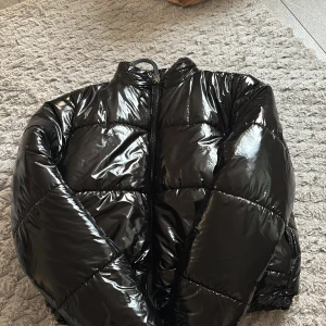 Svart glansig pufferjacka - Säljer en svart, glansig pufferjacka med hög krage och dragkedja framtill. Jackan har en oversized look och breda, vadderade paneler som ger en riktigt trendig vibe. Perfekt för kalla vinterdagar när du vill hålla dig varm och se cool ut.