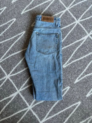 Blå jeans från Woodbird - Snygga ljusblå jeans från Woodbird med klassisk femficksdesign och rak passform. Jeansen har en tydlig logga på bakre linningen och är tillverkade i slitstark denim. Perfekta för en avslappnad och trendig stil. Storlek W28 L32. Obs knappen har lossnat men har satt dit en ny lite till sidan. Hör av er vid frågor eller lägg ett prisförslag!