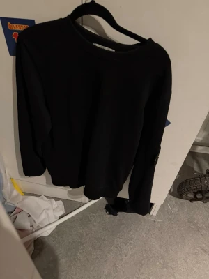 Svart sweatshirt från Stone Island - Säljer en svart sweatshirt från Stone Island med den klassiska loggan på vänster ärm. Tröjan har rund halsringning och långa ärmar. Perfekt för dig som gillar streetwear och enkel stil.