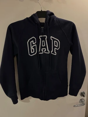 Mörkblå hoodie med dragkedja GAP - Mörkblå hoodie från GAP med vit broderad logga på bröstet. Tröjan har dragkedja framtill, två fickor och justerbar huva med snören. Tillverkad i mjukt sweatshirtmaterial, perfekt för en avslappnad stil.