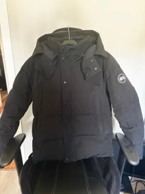 Svart dunjacka från Canada Goose - Säljer en svart dunjacka från Canada Goose med avtagbar huva och diskret logga på ärmen. Jackan har stora fickor framtill, dragkedja och knappar för extra skydd mot kyla. Perfekt för kalla vinterdagar och riktigt varm.