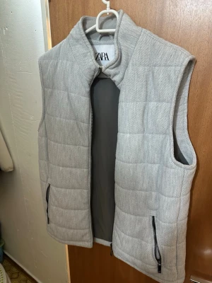 Ljusgrå dunväst från Zara - Stilren ljusgrå dunväst från Zara med quiltad design och hög krage. Västen har två praktiska fickor med dragkedja och en innerficka. Materialet är mjukt och lätt, perfekt för lager-på-lager. Passar till många olika outfits och ger en clean look.