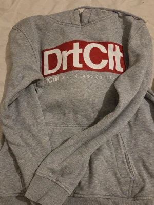 Grå DrtCult hoodie med tryck - Säljer en grå hoodie från DrtCult med stor vit och röd logga på bröstet och texten 'San Diego California' under. Klassisk känguruficka framtill och ribbade muddar vid ärmslut och nederkant. Perfekt för en avslappnad streetwear-look.