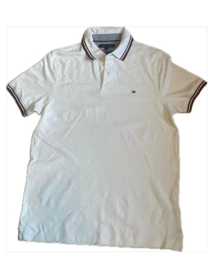 Vit piké från Tommy Hilfiger - Klassisk vit pikétröja från Tommy Hilfiger med krage och knappar framtill. Kragen har snygga ränder i rött, blått och vitt. Liten logga på bröstet och ärmsluten har matchande randiga detaljer. Perfekt till en clean och stilren look.